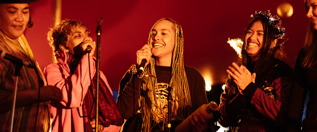 Four musicians standing on the Jambala stage. Two are singing. One is clapping and wearing fairy ears. The stage is red.